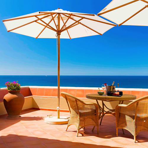 Eine Terrasse mit Korbstühlen und einem runden Tisch unter großen weißen Sonnenschirmen mit Blick auf den Ozean.