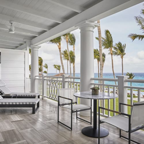Eine geräumige Außenterrasse mit Sonnenliegen, einem kleinen Tisch mit Stühlen, Blick auf einen Strand mit Palmen und dem Meer.
