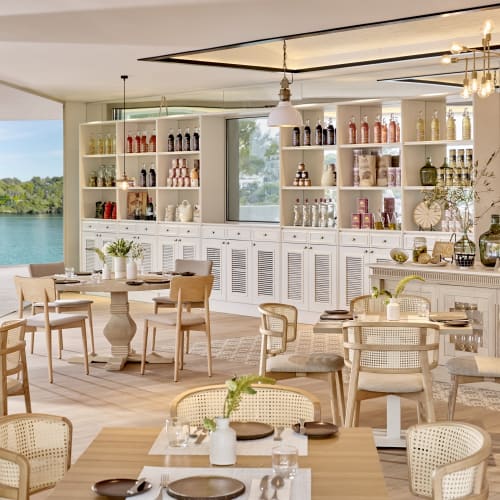 Interior of a bright restaurant with white shelves, tables, and chairs, overlooking a waterfront view.