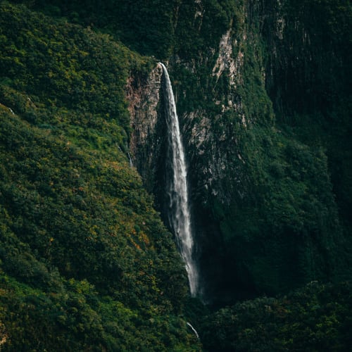 Tall waterfall cascading down a steep, lush green mountainside covered with dense vegetation.