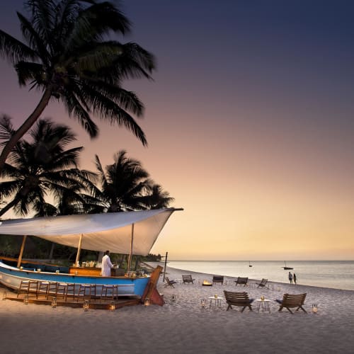 Ein Strandabschnitt bei Sonnenuntergang mit einem traditionellen Boot unter Palmen und Liegestühlen am Sandstrand.