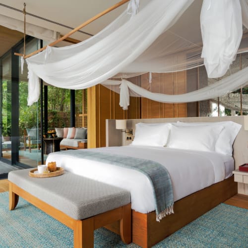Luxus Schlafzimmer mit einem Himmelbett und transparenten Vorhängen, mit Balkon und Outdoor-Möbeln, umgeben von üppigem Grün.