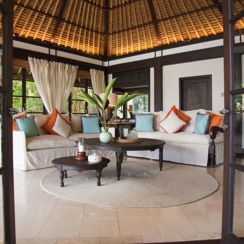 Elegantes Wohnzimmer mit weißen Sofas, bunten Kissen und großen Fenstern mit cremefarbenen Vorhängen in einer tropischen Umgebung.