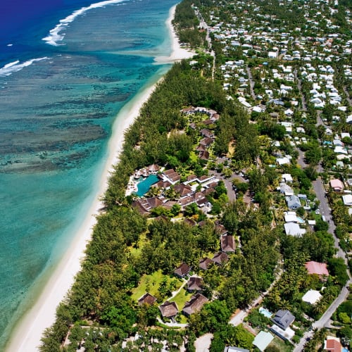 Aerial view of a coastal resort area with lush greenery, white sandy beaches, turquoise ocean waters, and numerous buildings including houses and vacation huts.