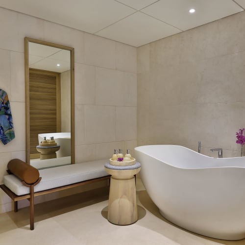 Modernes Badezimmer mit großer ovaler freistehender weißer Badewanne, einer Holzbank mit Polster, einem Beistelltisch mit Badeprodukten und einem bunten gemusterten Morgenmantel an der Wand unter einem rechteckigen Spiegel.