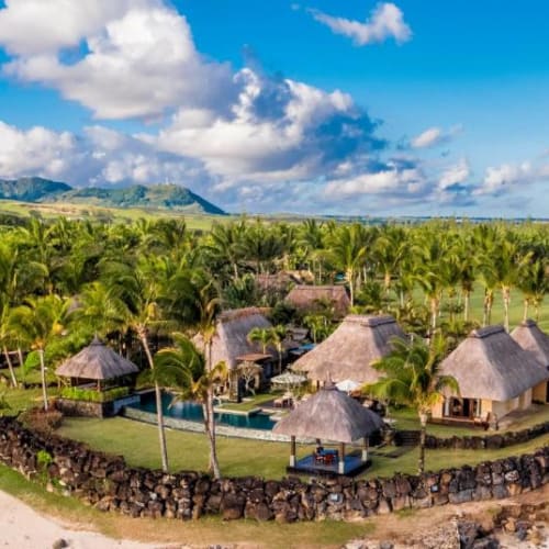 Aerial view of the Shanti Maurice Resort & Spa with thatched-roof villas surrounded by lush greenery, a swimming pool, and a sandy beach coastline.
