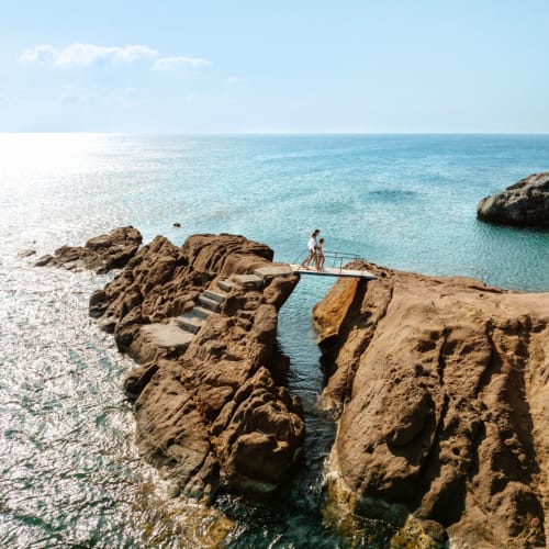 Zwei Personen gehen auf einer kleinen Brücke aus Felsen, die über das Wasser führt, mit dem Meer im Hintergrund und Felsen am Ufer.