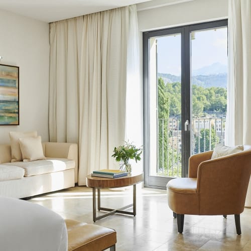 Helles Hotelzimmer mit großem Bett, beigem Sofa, braunen Sessel, rundem Holztisch mit Büchern und Blumen sowie großem Fenster mit cremefarbenen Vorhängen mit Blick auf Grünflächen und entfernte Hügel.