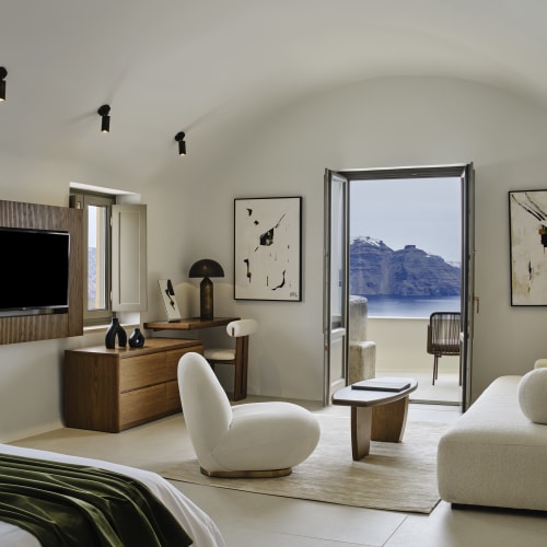Modern white living space with a curved ceiling, featuring a flat-screen TV, minimalist furniture including a white armchair and sofa, a small wooden coffee table, and an open glass door offering a view of a rocky coastline and blue sea.