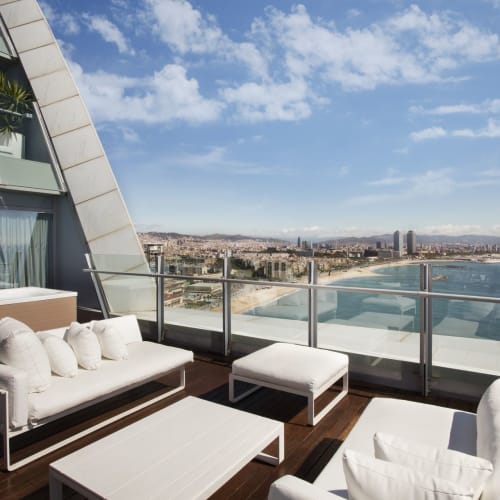 Luxurious terrace lounge with white outdoor furniture overlooking the sea and city skyline under a partly cloudy sky.