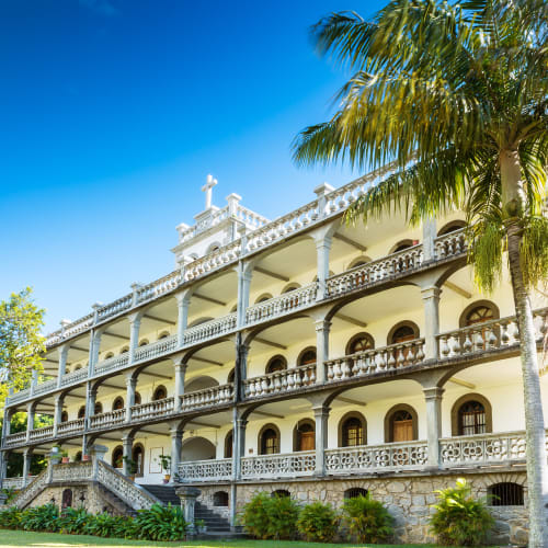 A multi-story white historic building with arches and balconies, surrounded by palm trees under a blue sky.