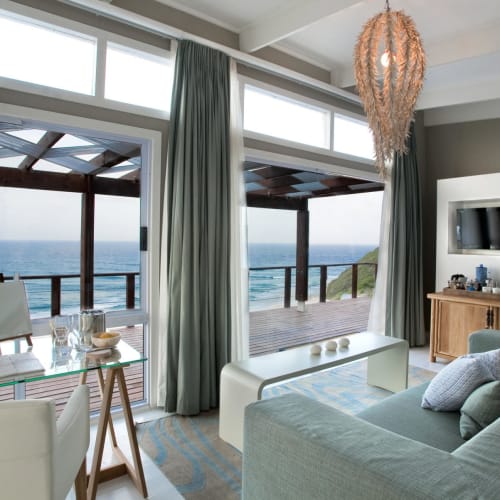 Luxuriöses Suite-Lounge mit Blick auf den Ozean, mit Glastisch, weißen Stühlen, einem Sofa mit Kissen und einem Fernseher.