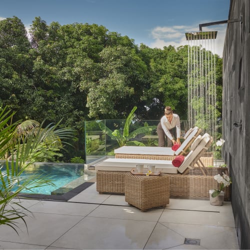 Eine Frau ordnet Handtücher auf Liegestühlen neben einem Außenpool mit moderner Außendusche auf einer gefliesten Terrasse umgeben von Bäumen und Grün.