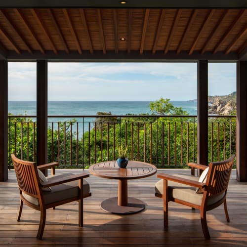 Eine Veranda mit einem runden Tisch und zwei Stühlen mit Blick auf eine grüne Landschaft und den Ozean.