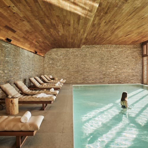 Ein ruhiger Innenpoolbereich mit einer Person im Wasser, umgeben von Holzarchitektur und Liegestühlen.