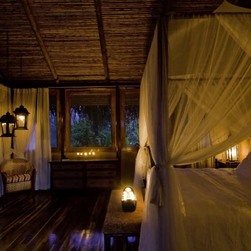 Cozy honeymoon suite interior with a four-poster bed and warm lighting, overlooking a tropical jungle through large windows.