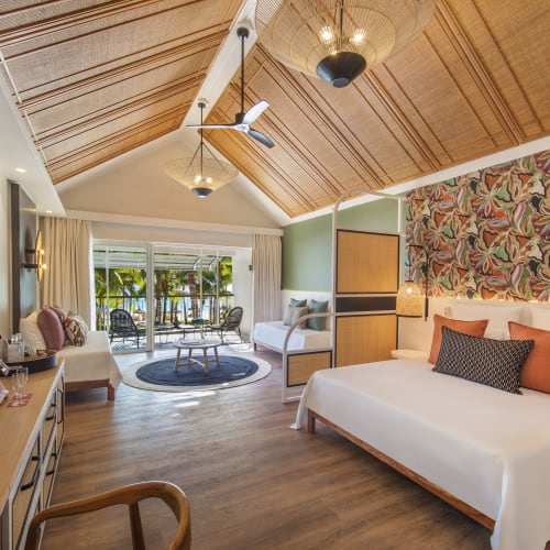Spacious hotel room with a high wooden ceiling, a large bed with decorative pillows, a sitting area with sofa and chairs, and an outdoor terrace overlooking palm trees and the beach.