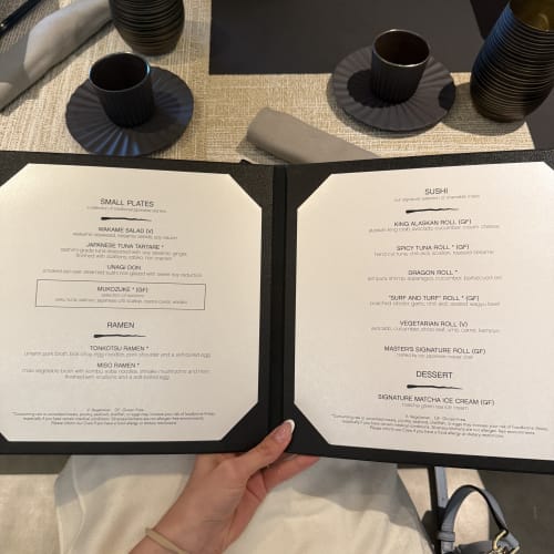 Eine Person hält in einem Restaurant eine aufgeklappte Speisekarte an einem gedeckten Tisch mit dunklen Tellern, Tassen und Servietten, die eine Auswahl an kleinen Gerichten, Ramen, Sushi und Desserts zeigt.