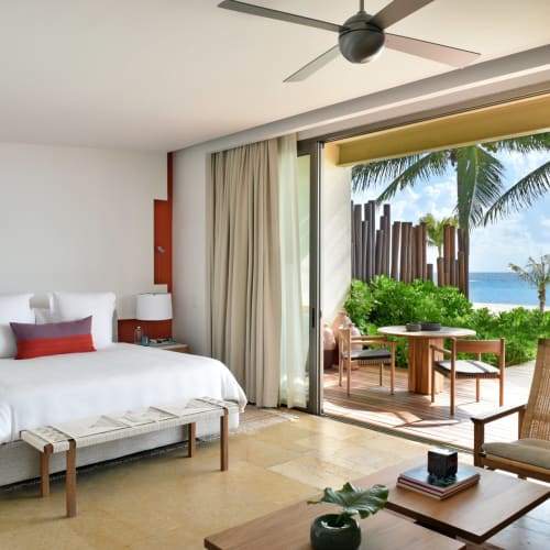 Ein luxuriöses Strandstudio mit großem Bett, angrenzender outdoor Sitzgelegenheit und Blick auf Palmen und den Ozean.