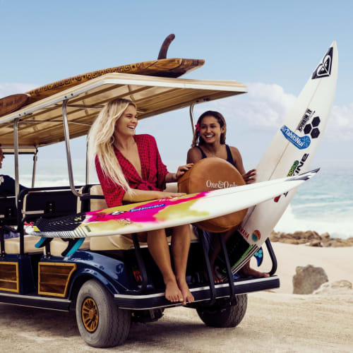 Zwei Frauen sitzen auf Surfboards auf einem Golfwagen am Strand, bereit zum Surfen.