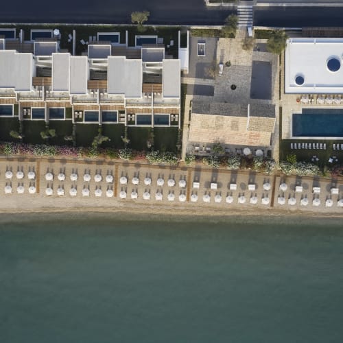 Aerial view of a beachfront resort with multiple villas, a swimming pool, and rows of sun loungers and umbrellas aligned on the sandy beach beside calm water.