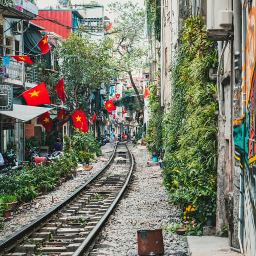 A narrow street in Hanoi, Vietnam, with a train track running through the middle, decorated with Vietnamese flags and lined with shops and vibrant graffiti.