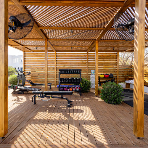 Outdoor gym area with wooden deck and pergola roof featuring exercise equipment including stationary bikes, kettlebells, dumbbells, and workout benches surrounded by plants and natural light.
