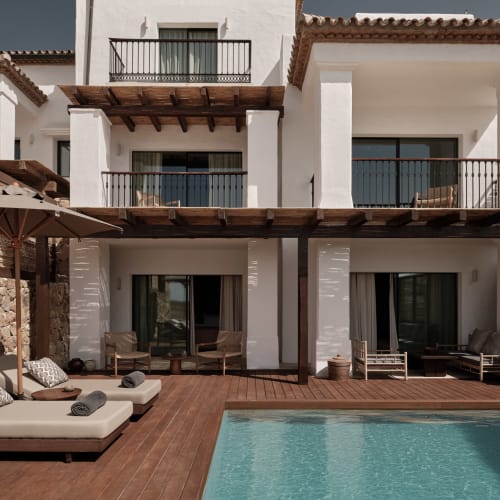Moderne weiße Villa mit privatem türkisfarbenem Schwimmbad, Holzdeck, Sonnenliegen mit Kissen und großen Sonnenschirmen in sonniger Außenanlage.