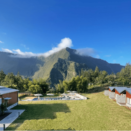 A scenic mountain landscape with lush greenery, clear blue sky, and several modern cabins and a swimming pool on a grassy area in the foreground.