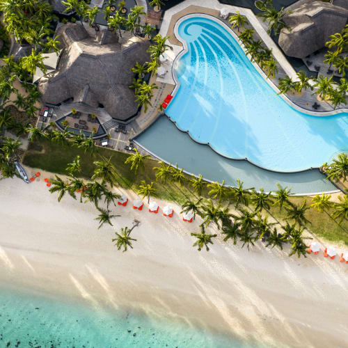 Aerial view of a tropical beach resort with a uniquely shaped swimming pool surrounded by palm trees and thatched-roof buildings, adjacent to a white sandy beach and turquoise ocean water.