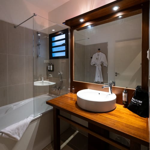 Modernes Badezimmer mit einer Holzarbeitsplatte, die ein rundes weißes Waschbecken hält, einem großen Spiegel mit integrierten Leuchten, einer Badewanne mit Glas-Duschabtrennung und einem weißen Handtuch am Wannenrand.