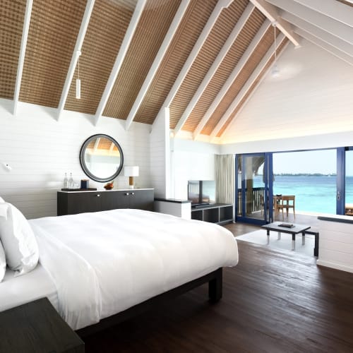 Water villa bedroom with large window offering a view of the ocean, decorated in white and dark wood furniture.