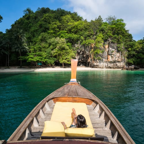 A woman relaxing on a yellow lounge chair at the bow of a longtail boat in a tropical bay with green cliffs and lush trees in the background.