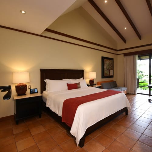 The bedroom features a large bed with white linens and a red runner, flanked by two bedside lamps, with a seating area and a balcony overlooking greenery.