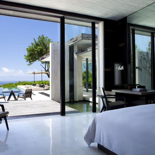 Luxury bedroom with a large bed, modern interior, and sliding glass doors opening to an outdoor patio with seating and views of greenery and the ocean.
