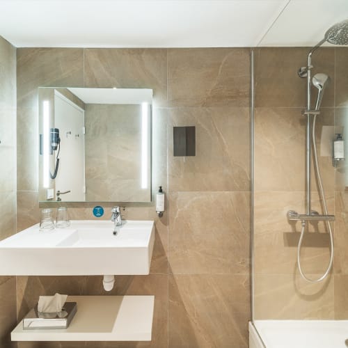 Modernes Badezimmer mit beige gefliesten Wänden, weißem Waschbecken, beleuchtetem Spiegel, wandmontiertem Haartrockner, Glaskabine mit Regendusche und einem kleinen Regal mit Handtüchern unter dem Waschbecken.
