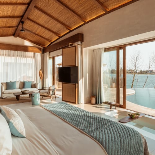 Luxuriöses Schlafzimmer in einer Flusspoolvilla mit großem Bett, Sitzbereich, Flachbildfernseher und Schiebetüren, die zu einem Infinity-Pool mit Blick auf einen Fluss und kahle Bäume führen.