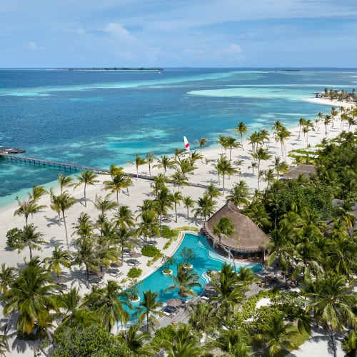 Luftaufnahme eines tropischen Inselresorts mit weißem Sandstrand, Palmen, Wasserbungalows und einem türkisfarbenen Swimmingpool, umgeben von üppigem Grün neben dem Ozean.