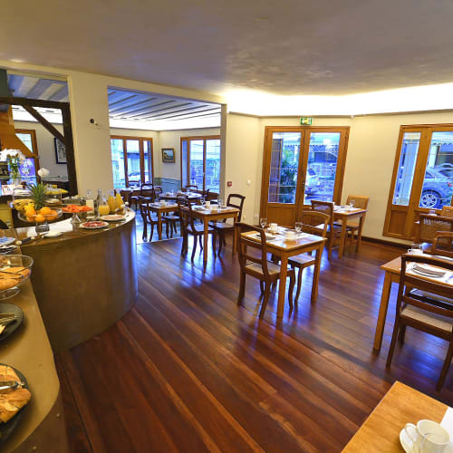 Gemütlicher Frühstücksraum mit Holzboden, mehreren Holztischen und Stühlen, die mit Tassen und Tellern gedeckt sind, sowie einem Buffet mit Gebäck und Obst in einem warm beleuchteten Speiseraum.