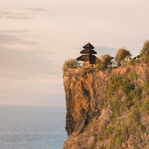 A traditional Balinese temple with tiered roofs is perched on the edge of a high cliff overlooking the ocean at sunset.