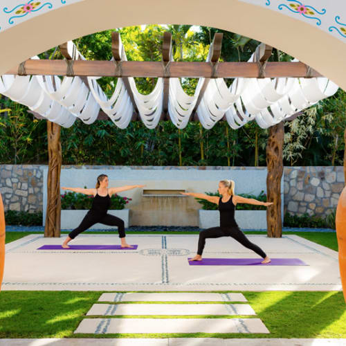 Zwei Frauen, die Yoga im Freien unter einer dekorierten Pergola praktizieren, mit üppigem Grün und großen Tonkrügen in der Nähe.