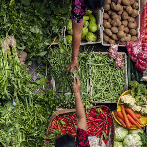 A vendor at a busy outdoor market selling fresh vegetables and greens.