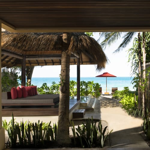 A tropical beachfront patio with a thatched-roof daybed adorned with red cushions, palm trees, and a sandy pathway leading to a red beach umbrella by the ocean.