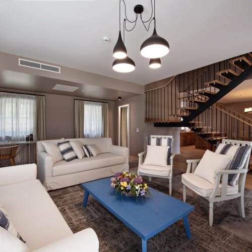 Elegant living room with white sofas and armchairs, a blue coffee table with a floral arrangement, and a wooden staircase in a modern hotel interior.