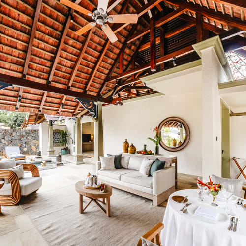 Luxuriöse Outdoor-Villa-Suite mit Holzdach, Deckenventilator, weißem Sofa, runden Sesseln, rundem Couchtisch und einem für zwei Personen gedeckten Tisch neben einem Pool mit Liegestühlen.