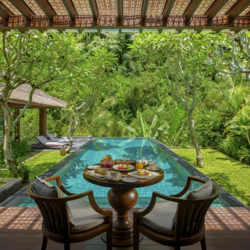 A luxurious outdoor terrace with a round dining table set for breakfast, overlooking a swimming pool and lush tropical greenery.