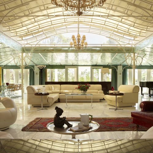 Luxurious living room with modern white sofas, a red armchair, a grand piano, and large windows overlooking a garden.