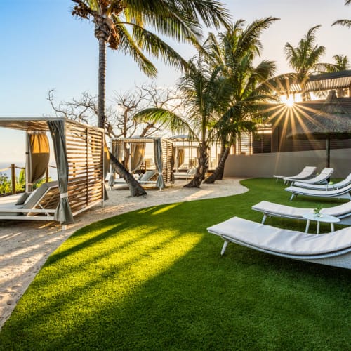 Ein Strandentspannungsbereich mit weißen Liegestühlen auf grünem Rasen und Baldachinbetten am Sandstrand unter Palmen bei Sonnenuntergang.