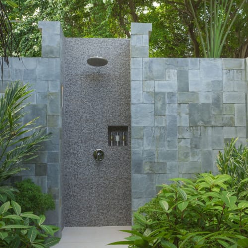 An outdoor shower area with a stone wall, surrounded by lush green plants and trees.