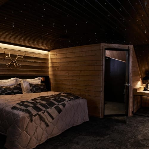 Gemütliches Schlafzimmer im Dachgeschoss mit Holzverkleidung an Wänden und Decke, einem ordentlich gemachten Bett mit gemusterten Bettwäsche und Kissen, einem kleinen Schreibtisch mit Lampe und einem Stuhl bei sanfter Umgebungsbeleuchtung.